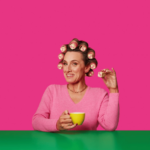 Woman sat in front of a pink background, holding a battenberg cake and using battenburg cakes as hair rollers | The Fife Arms