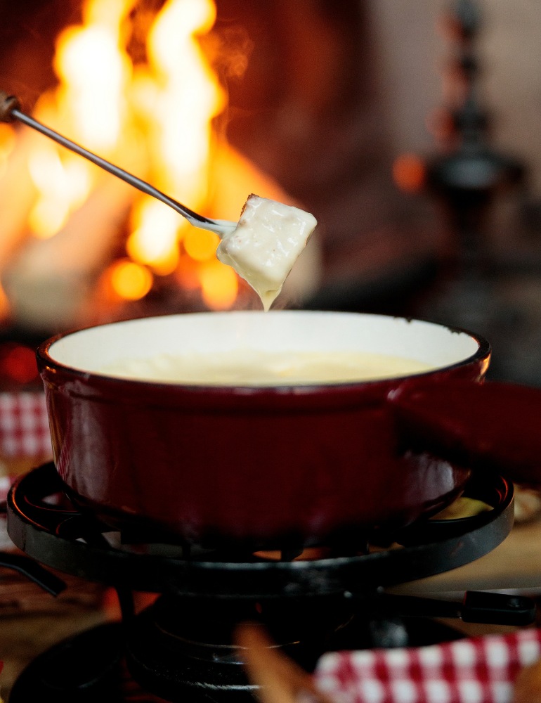 A piece of bread on a skewer being dipped in cheese fondue with a fire roaring in the background.