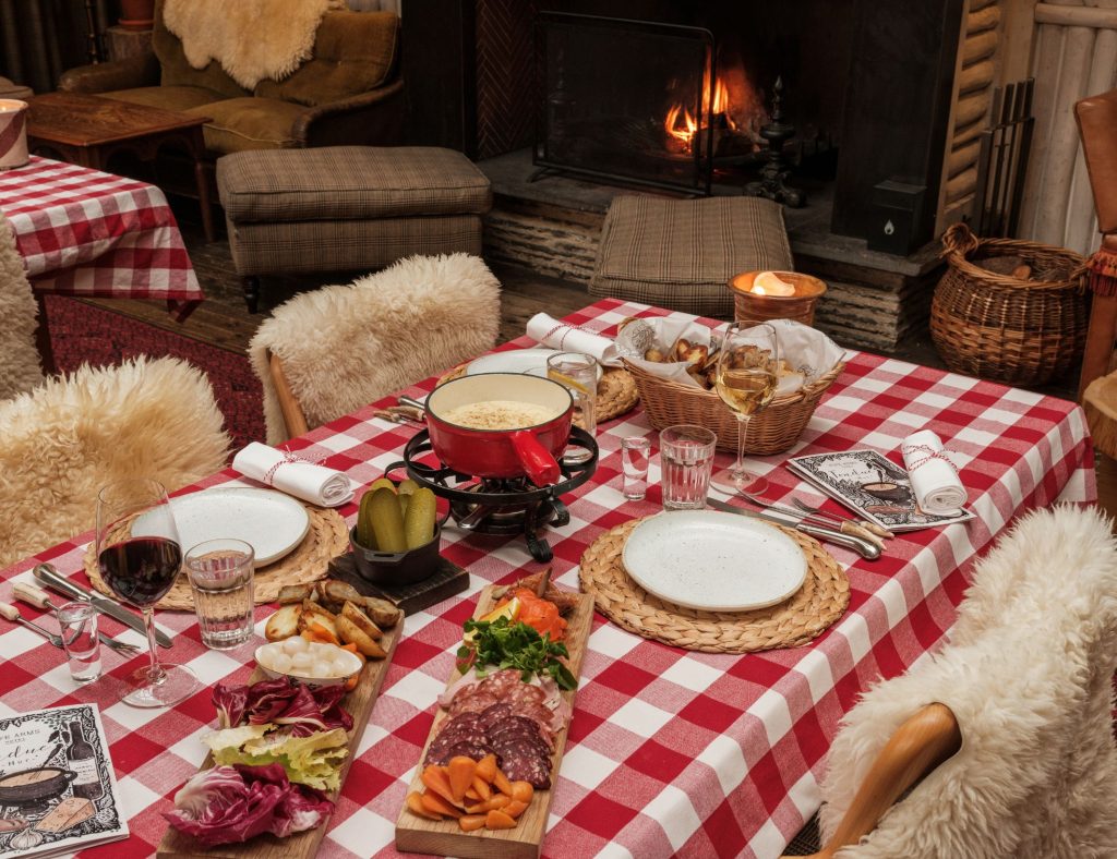 A dinner table laid for fondue with charcuterie, sheepskin rugs on the chairs and a fire burning in a fireplace