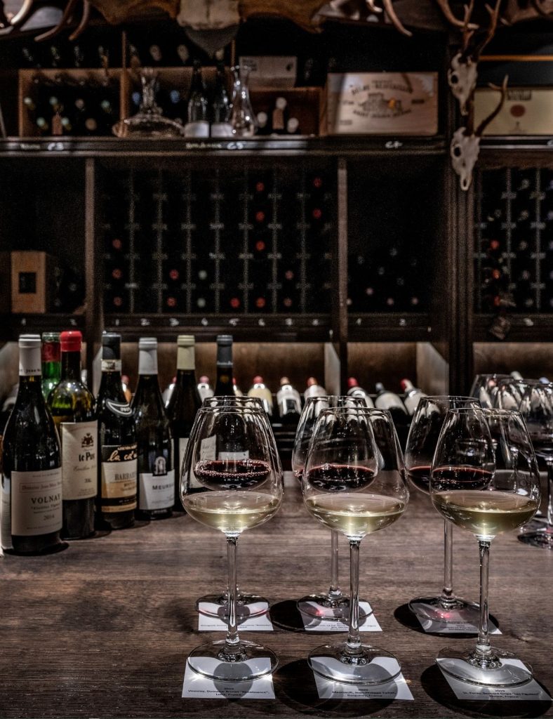 Wine glasses arranged on a wooden counter in front of shelves filled with bottles in the cellar for wine tasting.