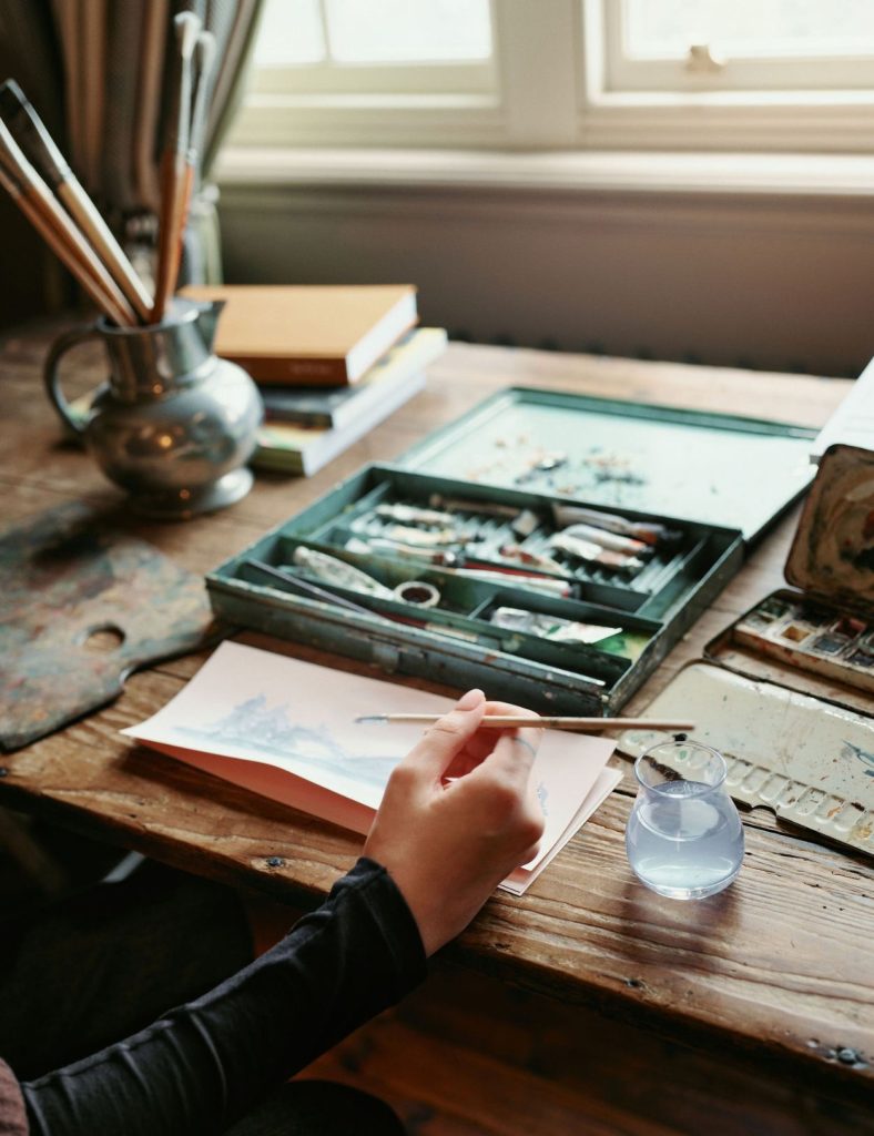 An artist using watercolours at a table in front of a window with a box of paints on and a green jug filled with paintbrushes.
