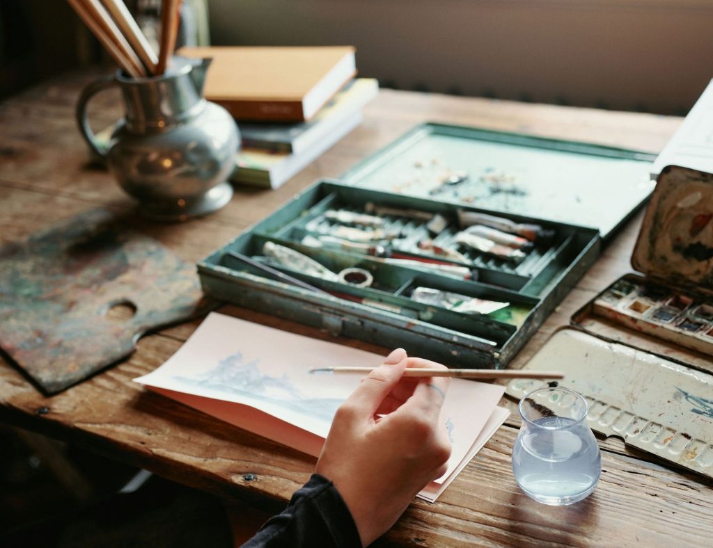An artist using watercolours at a table in front of a window with a box of paints on and a green jug filled with paintbrushes.