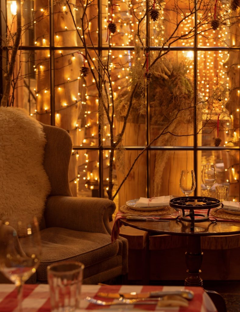 A table set up for fondue with an armchair in front of a glass window overlooking a courtyard with twinkling fairy lights