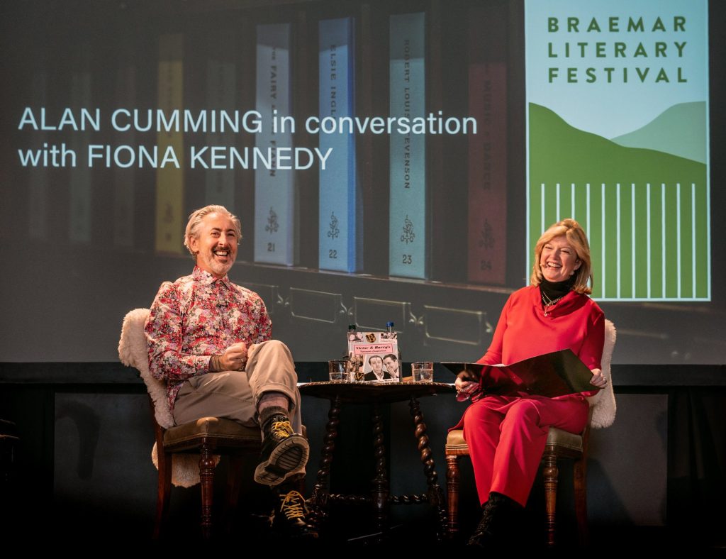 Alan Cumming and Fiona Kennedy sat on a stage with a screen behind them, both are laughing.