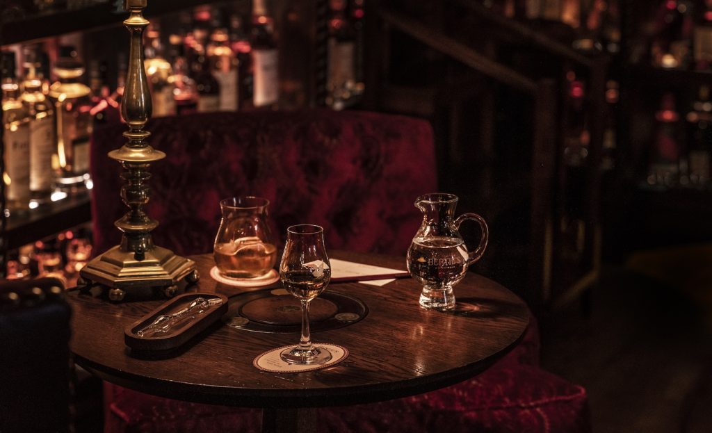 A dimly lit table with a tasting whisky glass, a water jug, a lamp, and bottles of whisky in the background.