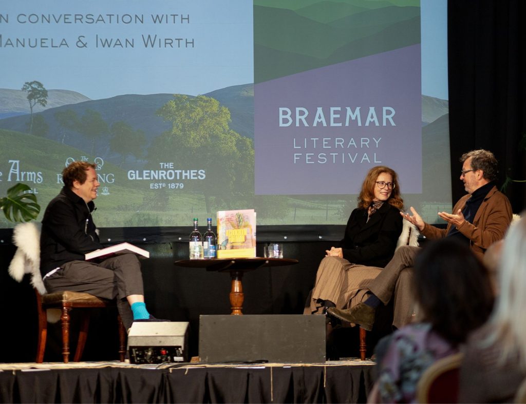 Three people talking on stage as part of the literary festival, with a book on a table in between them