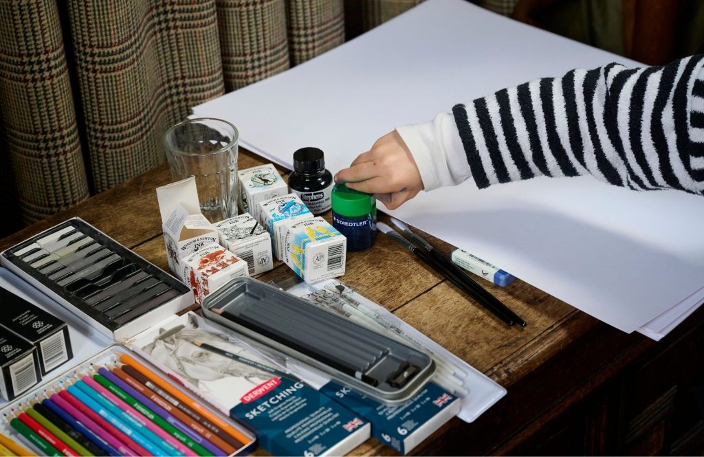 A child reaching for paintbrushes and paint on a table of art supplies.