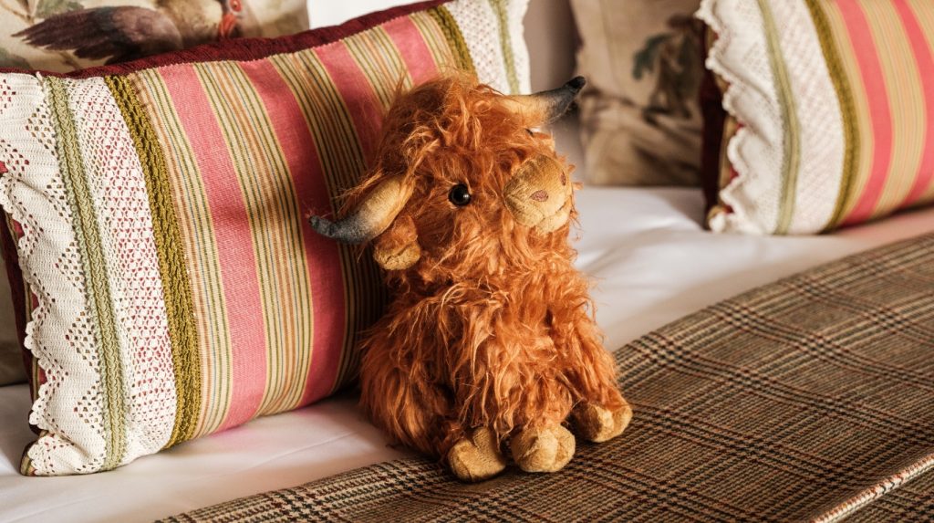 A soft Highland cow toy sitting on a bed in a family suite with patterned cushions and a tartan blanket.