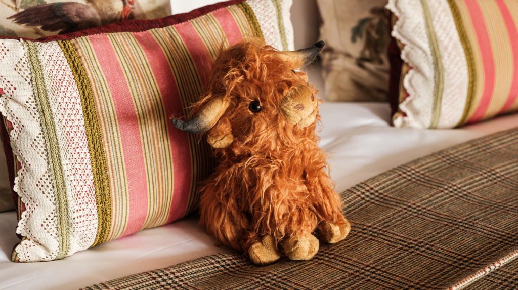 A soft Highland cow toy sitting on a bed in a family suite with patterned cushions and a tartan blanket.