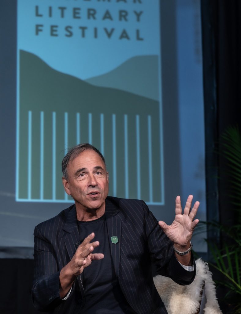 A close up of speaker Anthony Horowitz dressed in a stripe jacket, addressing a crowd whilst seated on a chair.