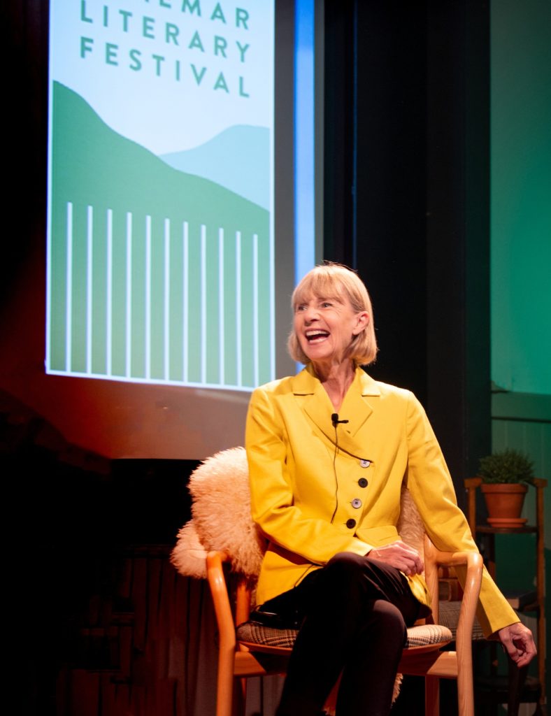 A close up of speaker Kate Mosse, dressed in a bright yellow jacket, smiling our to the crowd whilst seated on a chair.