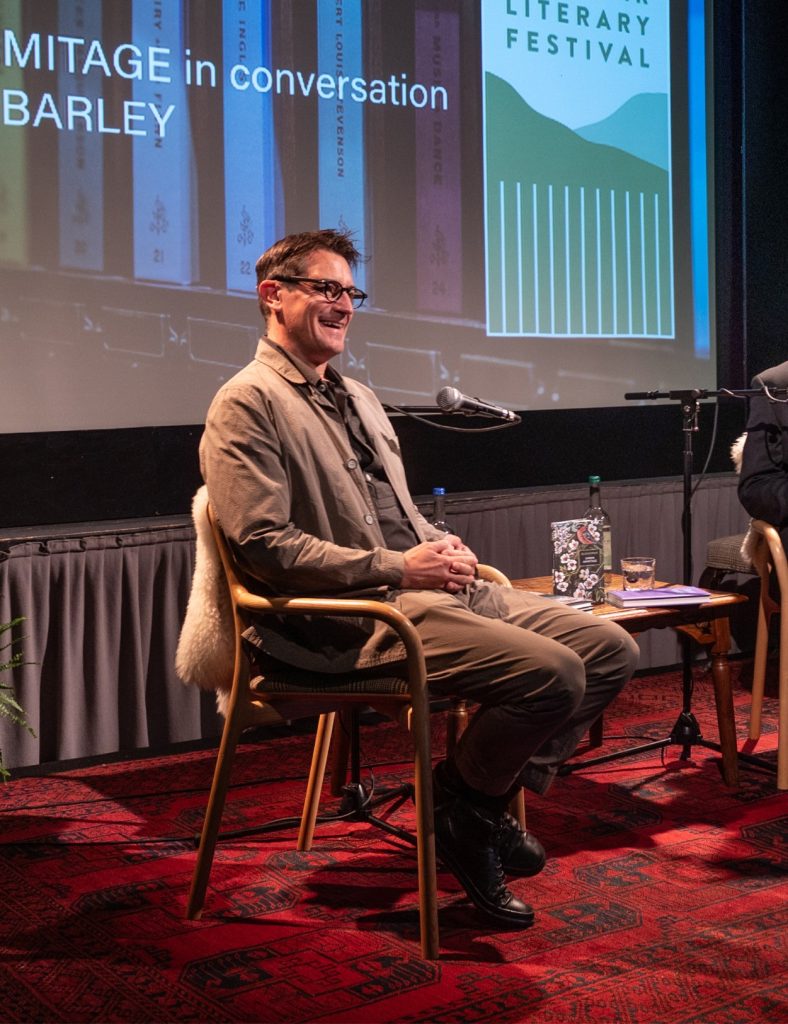 A close up of a speaker sitting on a stage covered in red carpet, looking out to the audience and holding a bottle of water.