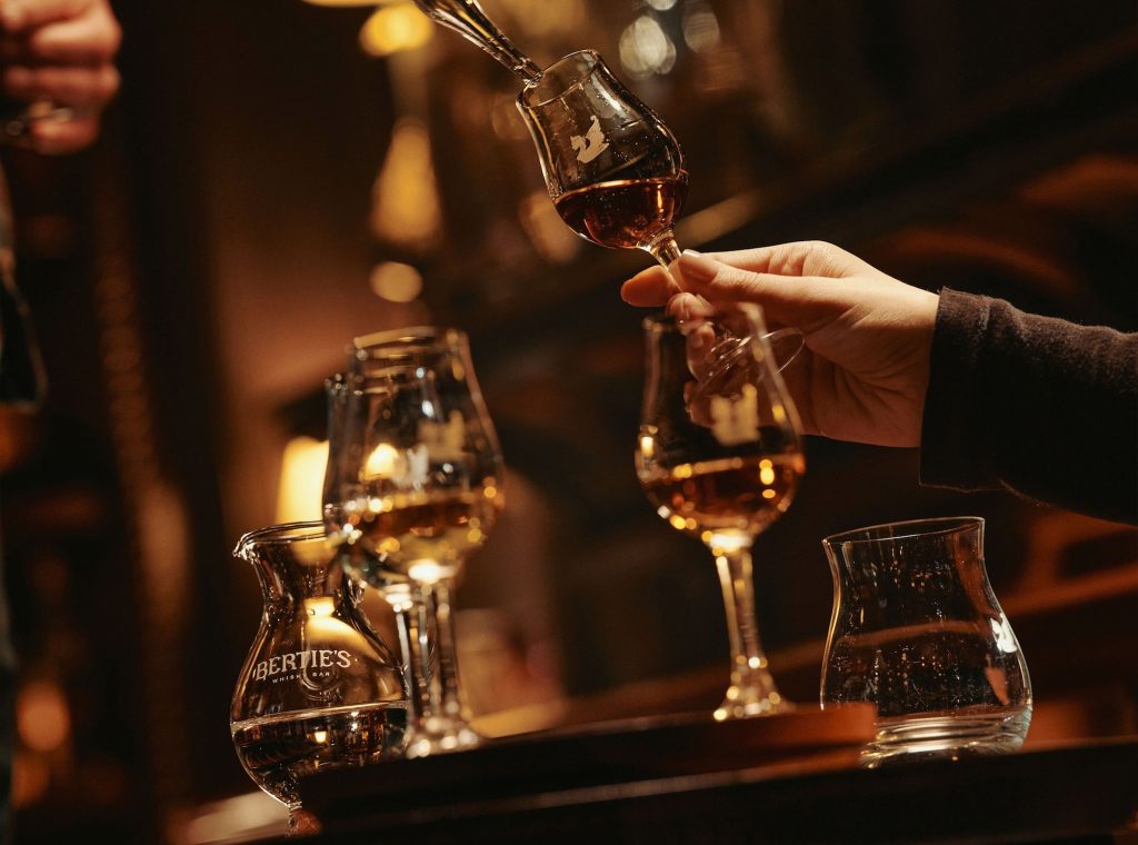 Close-up of a hand holding a tasting whisky glass as the spirit is poured, with several filled glasses on the bar.