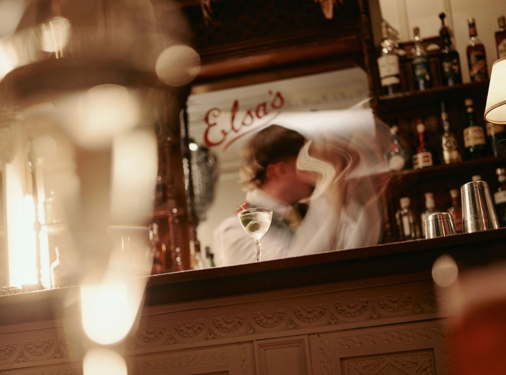 Bartender shaking a cocktail behind the bar at Elsa's Cocktail Bar with bottles on shelves and a martini glass in the foreground.