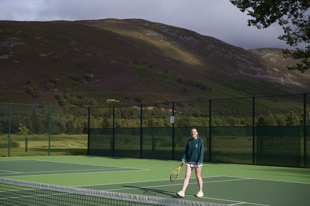 Woman in tennis gear holding a tennis racket and smiling on a tennis court with mountains in the background.