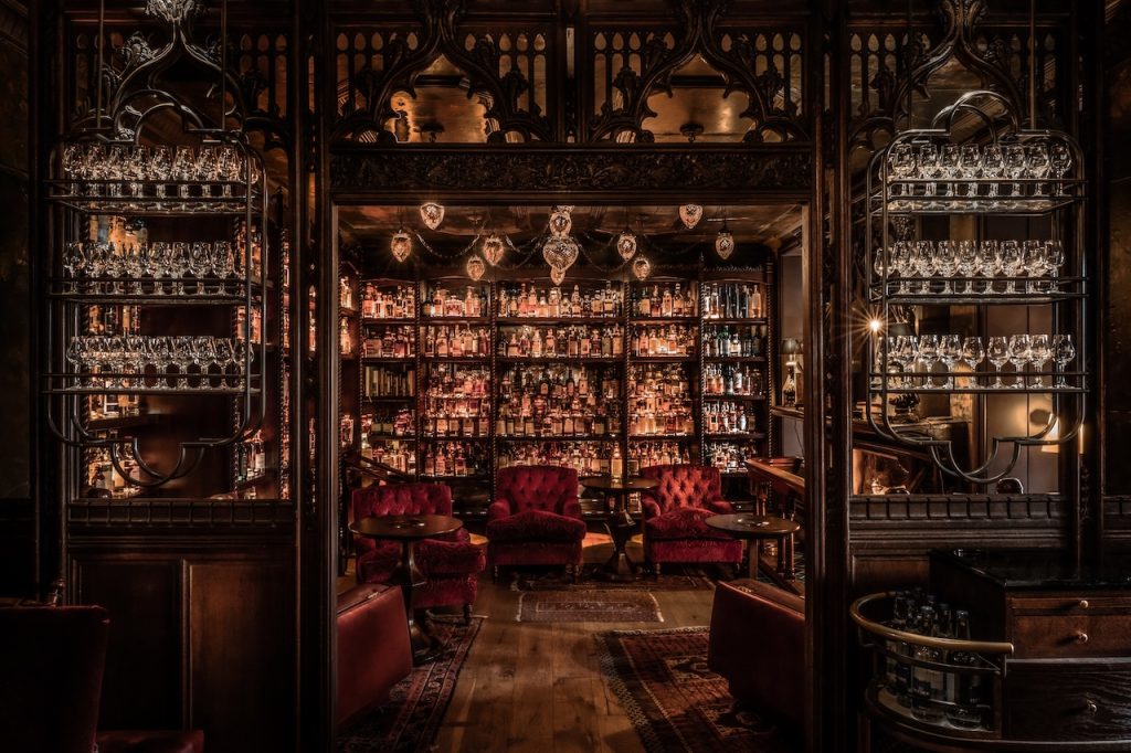 Wide shot of Bertie's Whisky Bar with red velvet chairs, ornate dark wood panels, and shelves filled with glassware and bottles.