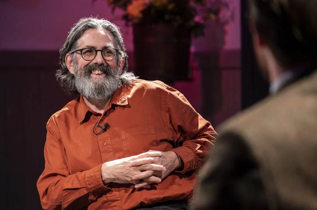 Man with beard in orange shirt smiling during a presentation