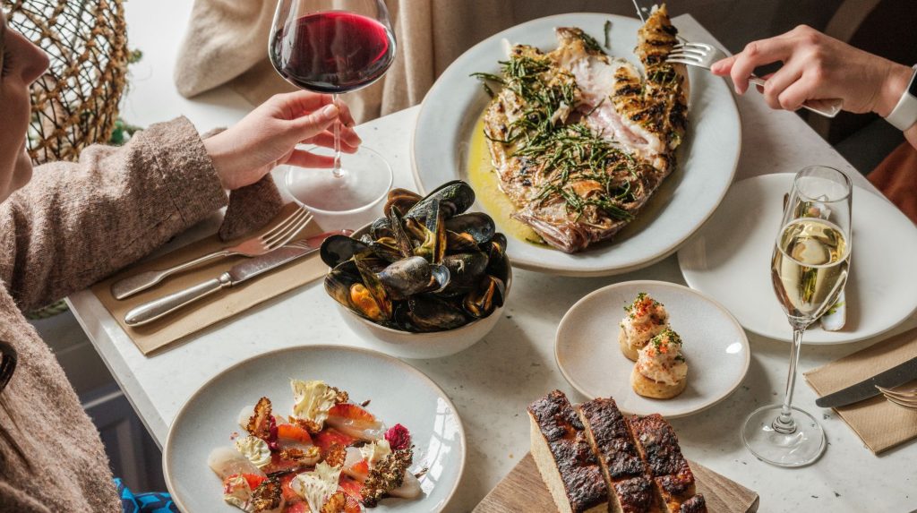 People sitting at a table at Fish Shop restaurant laid with a whole roasted fish, bowl of mussels, and glasses of red and sparkling wine.