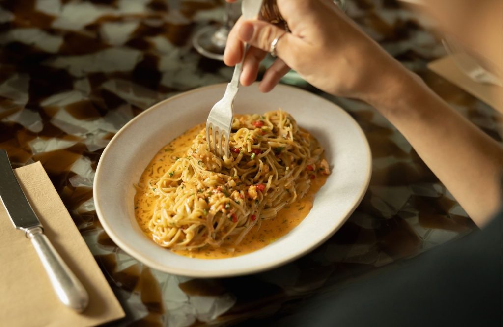 A hand twirls a forkful of spaghetti coated in a creamy, orange‑tinted sauce on a plate, set on a patterned table with cutlery nearby.
