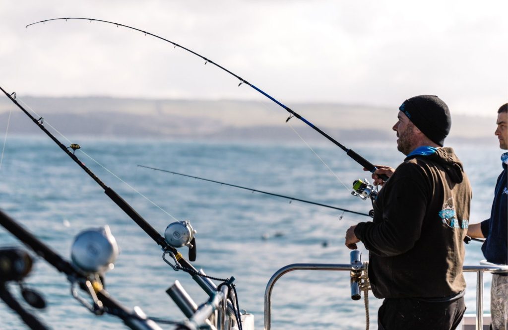 A fisherman stands at the rail of a boat holding a fishing rod over open water, with additional rods mounted nearby and distant hills visible across the sea.