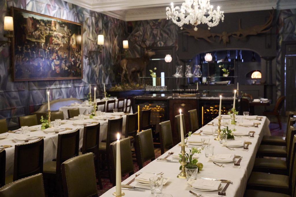 Elegant dining room with long candlelit tables, upholstered chairs, patterned walls, and a chandelier.