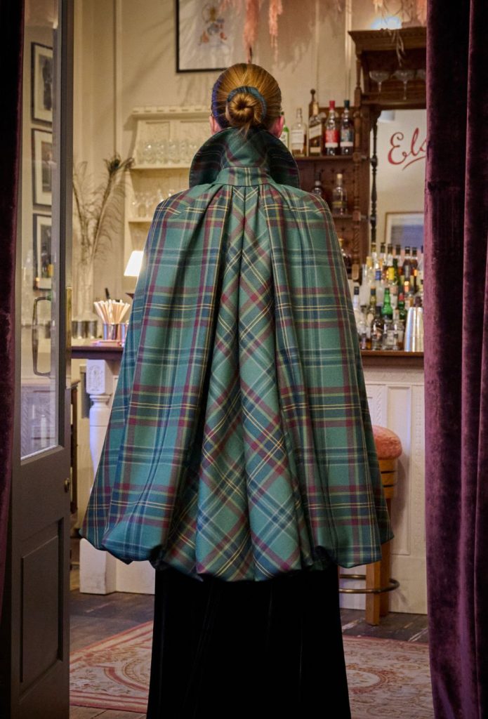 A woman stands inside a bar area wearing a long green tartan cape with a high collar, viewed from behind.