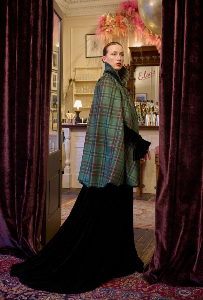 A woman wearing a tartan cape over a long black evening dress, standing in the doorway to a cocktail bar.