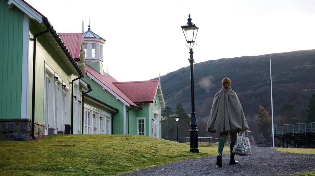 A woman wearing a tweek cape walks along a path beside pale green buildings with red roofs, with hills rising in the background.