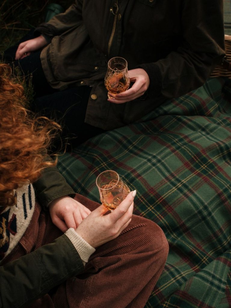 Two hands holding glasses of whisky above a green tartan picnic blanket