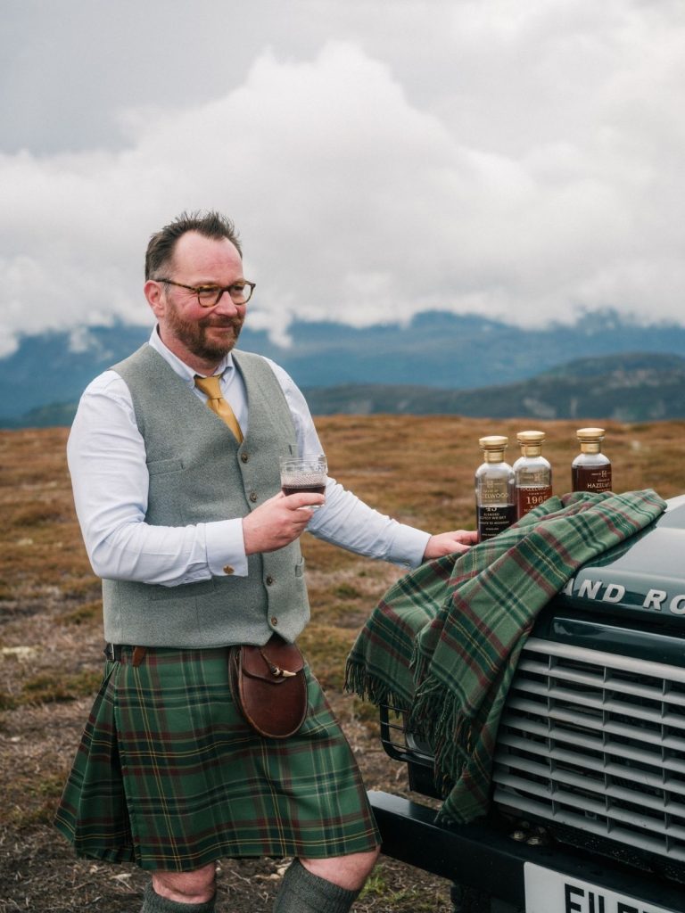 A man wearing a kilt holding a whisky glass beside a Land Rover, with bottles of whisky on its bonnet