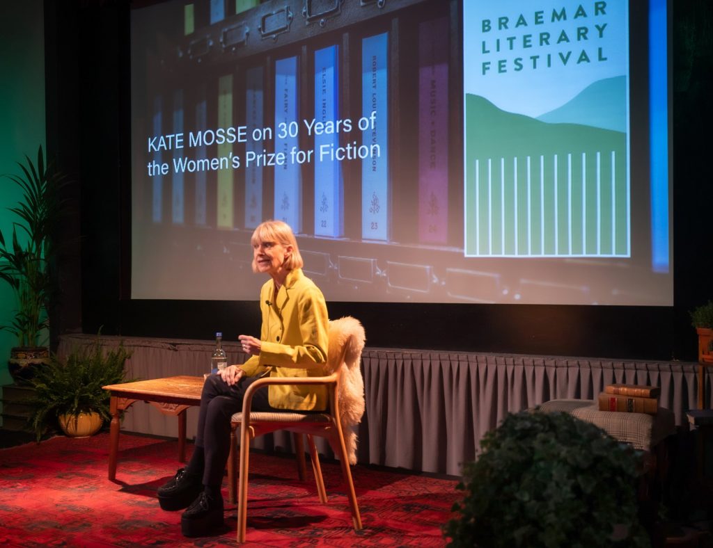A speaker called Kate Mosse sits on stage with a large screen behind them displaying the title ‘Kate Mosse on 30 Years of the Women's Prize for Fiction'.