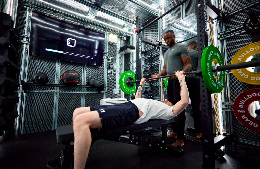 Person lifting a barbell on a bench press with a spotter in the compact PT pod gym space with weight plates and a wall‑mounted screen.