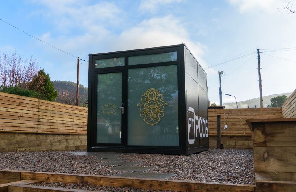Exterior of cube‑shaped PT pod gym unit with glass doors and a gold Fife Arms logo, set on gravel with wooden fencing around it.