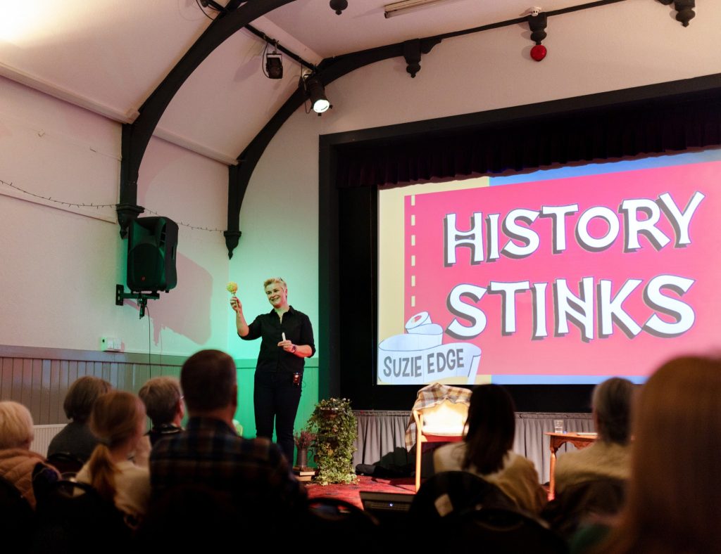 A speaker stands on stage in a hall holding an object while addressing an audience, with a large screen behind them displaying the title ‘History Stinks’ by Suzie Edge.
