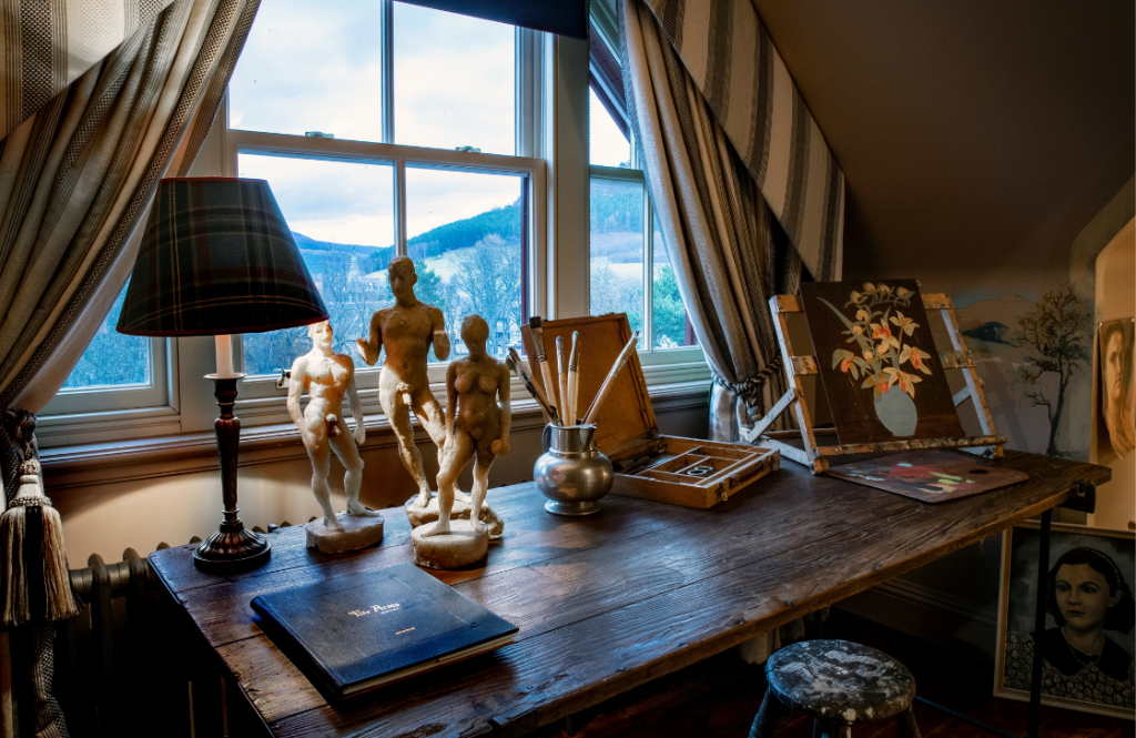 Wooden working desk in the Artist's Studio with art materials and sculpted figures by a window with rural hill views.