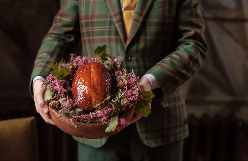 A person wearing tweed holding a roasted duck on a nest of heather.