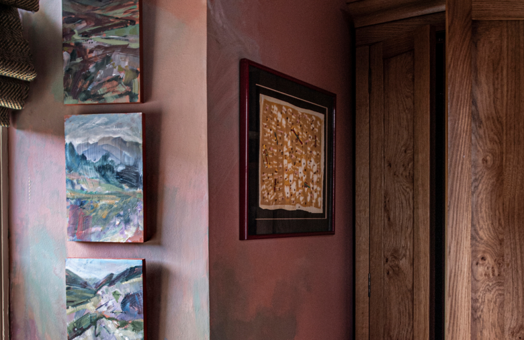 Wall with three contemporary paintings of mountains and a framed abstract artwork beside a wooden door in the Hill croft room.