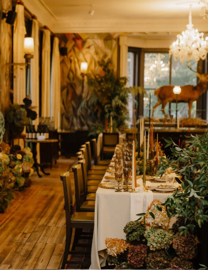 A long, elegantly set wedding dining table with tall candles and glassware in room filled with plants, warm lighting, and a taxidermy deer displayed near large windows.