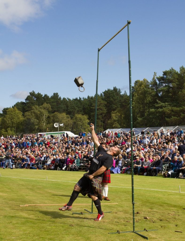 A competitor in a kilt throws a heavy weight upward in a outdoors games event, with a large crowd watching from stands on a sunny day.