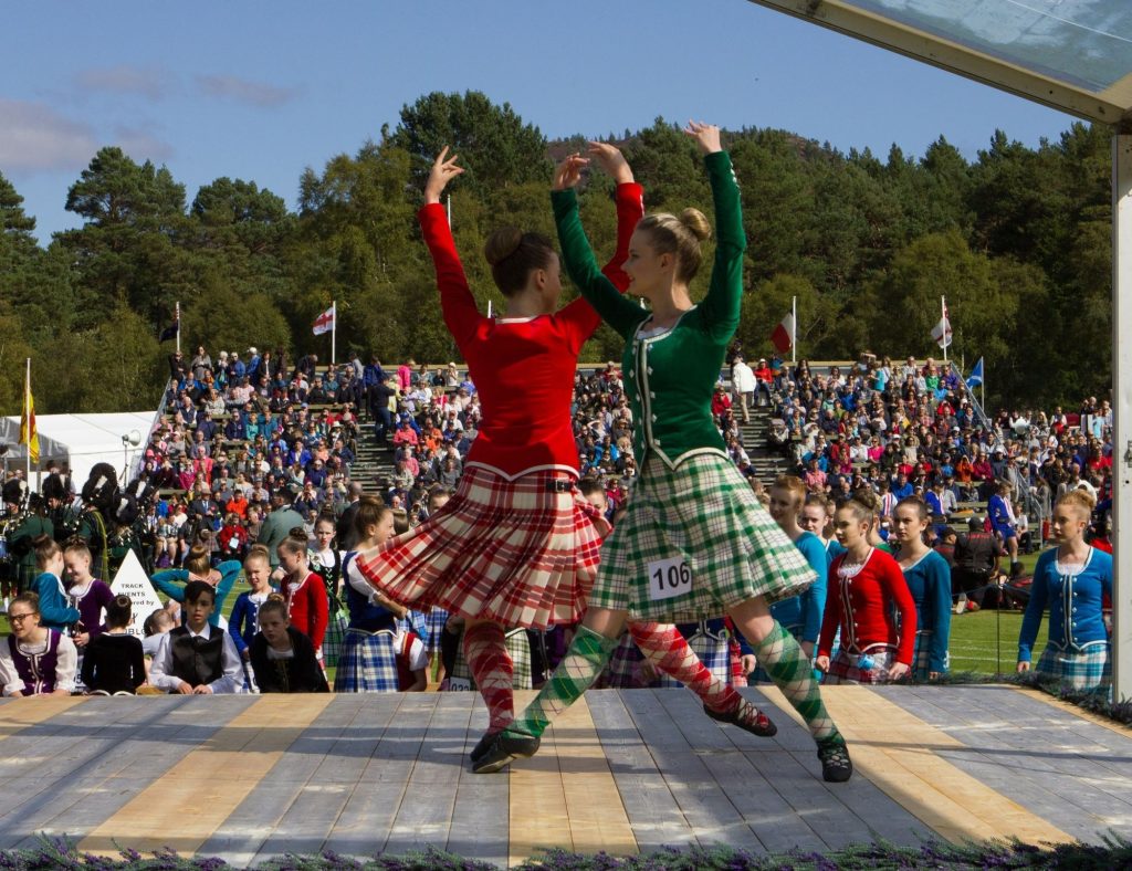 Highland dancers dressed in green and red dance on a wooden stage outside in front of spectators.