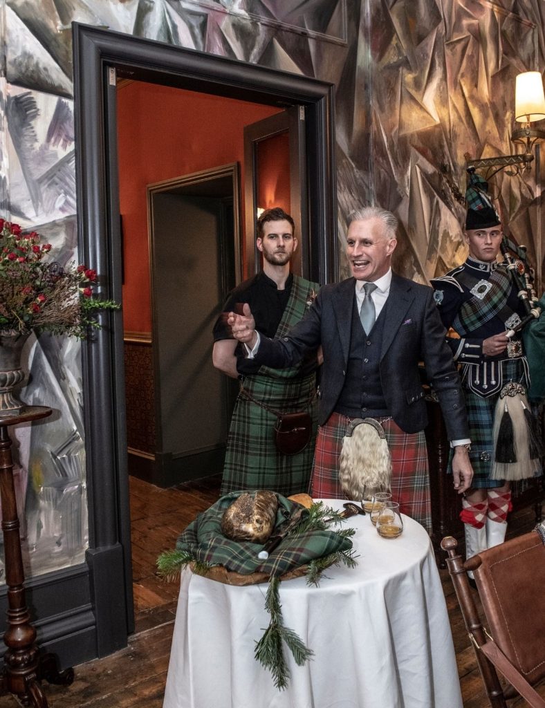 A man in a kilt addresses a haggis that sits on a table covered with a white cloth, while two men stand behind him.