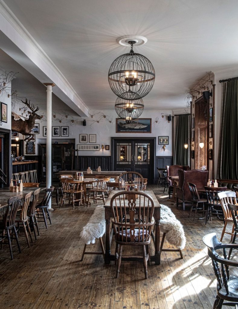 Empty interior of the Flying Stag pub with wooden tables and chairs, large spherical chandeliers, art filled walls, and warm natural light filling the room.