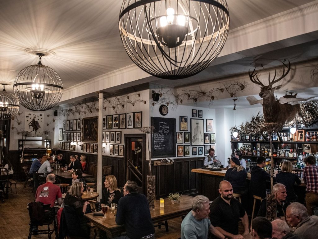 The lively interior of the Flying Stag pub with people seated at wooden tables and at the bar, surrounded by framed artwork, warm lighting, and large decorative chandeliers.