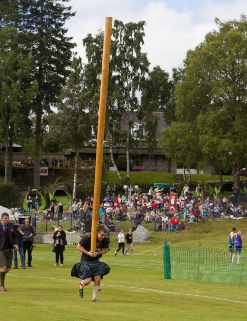 A heavy weight competitor in a kilt carries a large wooden caber across a grassy field during a games event in the Scottish Highlands, with spectators watching from the stands.