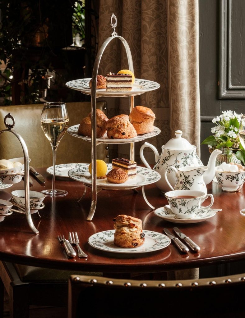 An afternoon tea on a brown wooden table in front of a window. Cakes and sandwiches are on a silver three-tiered cake stand on the table next to a tea pot.