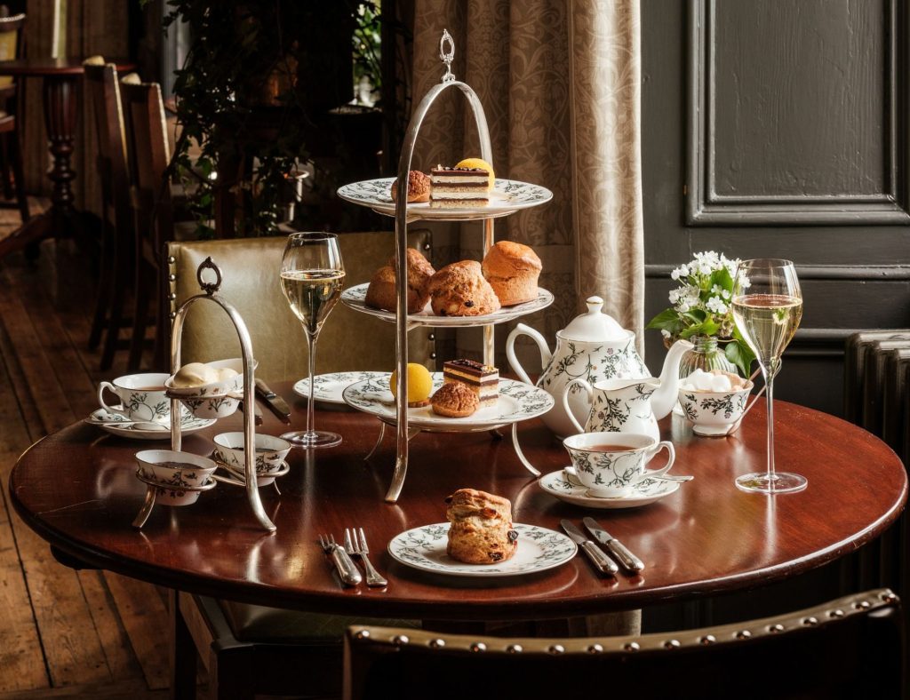 An afternoon tea on a brown wooden table in front of a window. Cakes and sandwiches are on a silver three-tiered cake stand on the table next to a tea pot.