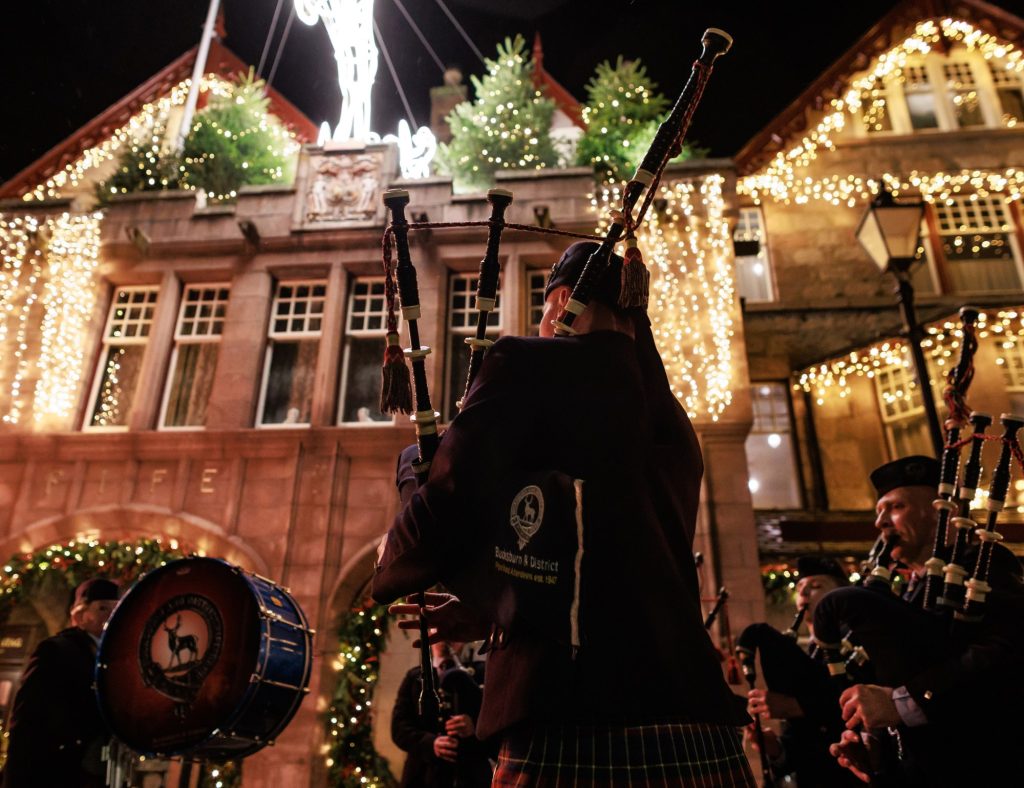 bagpipers playing outside of the Fife Arms Hotel entrance at night, the hotel is decorated with Christmas decorations and lights.
