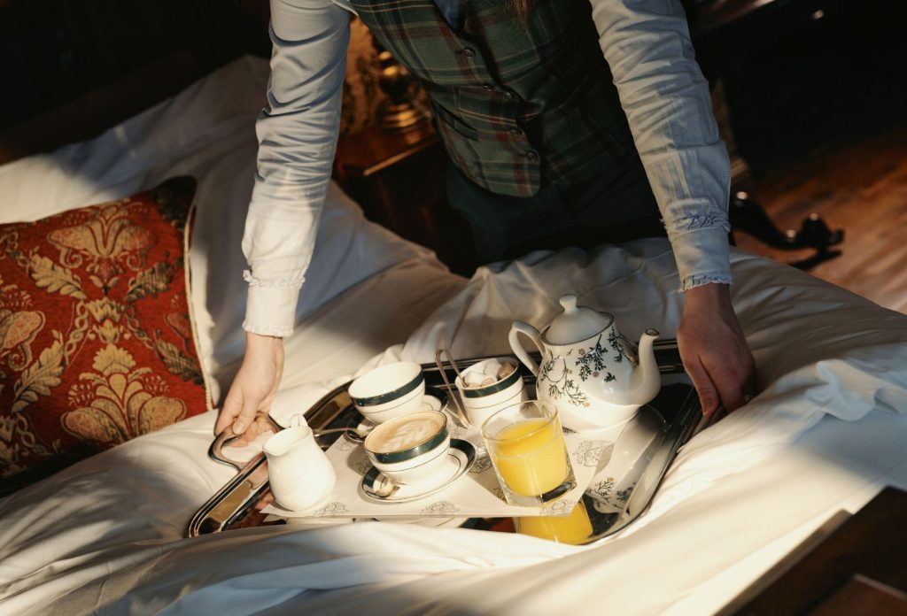 A silver breakfast tray with tea and coffee on being placed onto a hotel bed.