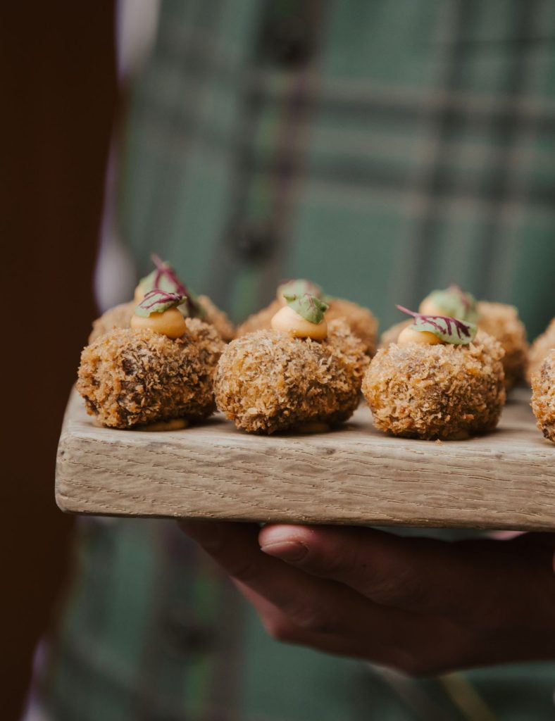 Canapes on a wooden board being held by a waiter wearing a green tartan waistcoat.
