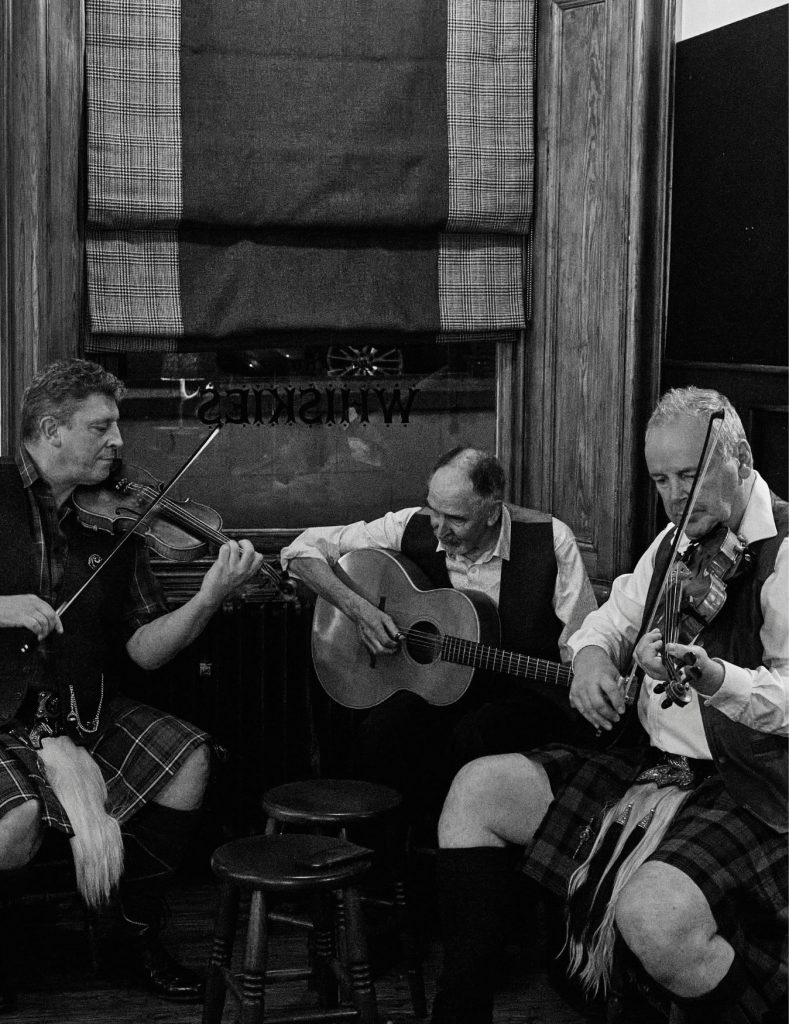 Three male Scottish folk musicians sat playing their string instruments in a pub.